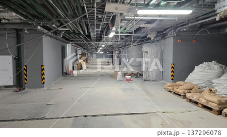 Unfinished corridor in a construction site featuring exposed pipes and electrical wiring overhead, with construction materials like bags and pallets arranged along the floor in a spacious area Unfinished corridor in a construction site featuring exposed pipes and electrical wiring overhead, with construction materials like bags and pallets arranged along the floor in a spacious area 137296078