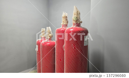 Four red fire extinguishers encased in protective netting stand against gray wall within storage area, highlighting essential safety equipment for fire prevention and emergency response measures Four red fire extinguishers encased in protective netting stand against gray wall within storage area, highlighting essential safety equipment for fire prevention and emergency response measures 137296080
