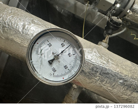 Pressure gauge attached to insulated metal piping in an industrial environment, showing readings from 0 to 100, with visible details of the dial and surrounding mechanical components Pressure gauge attached to insulated metal piping in an industrial environment, showing readings from 0 to 100, with visible details of the dial and surrounding mechanical components 137296082