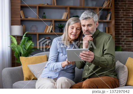Senior couple on a cozy living room sofa sharing a tablet, smiling and browsing the internet together, enjoying technology, connection and relaxed retirement moments Senior couple on a cozy living room sofa sharing a tablet, smiling and browsing the internet together, enjoying technology, connection and relaxed retirement moments 137297042