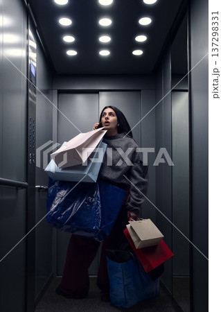 Surprised woman talking on the phone holding multiple shopping bags inside modern elevator. Surprised woman talking on the phone holding multiple shopping bags inside modern elevator. 137298331