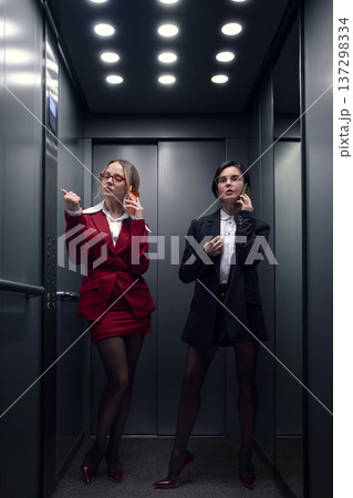 Two stylish businesswomen talking on smartphones in office elevator interior. 137298334