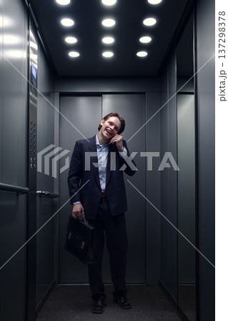 Young businessman smiling while talking on smartphone in modern office elevator. 137298378