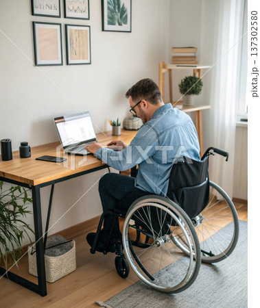 Man in wheelchair working on laptop at wooden desk in bright home office with plants and framed pictures on wall 137302580