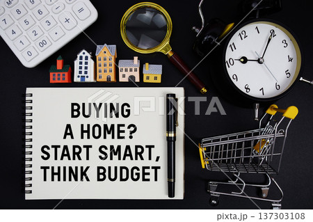Notepad with the phrase "Buying A Home? Start Smart, Think Budget" surrounded by a pen, mini trolley shopping cart, magnifying glass, clock, and house model 137303108