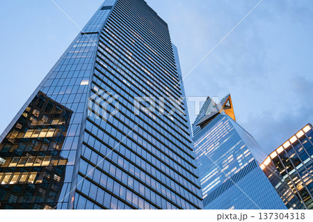 A tall glass skyscraper stands against a blue evening sky in Manhattan. Office lights illuminate the modern tower, symbolizing finance and business in New York City. A tall glass skyscraper stands against a blue evening sky in Manhattan. Office lights illuminate the modern tower, symbolizing finance and business in New York City. 137304318