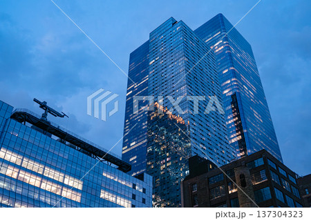 A modern glass skyscraper reflects the blue evening sky in Manhattan. Office lights glow inside the tower, highlighting corporate life in New York City. A modern glass skyscraper reflects the blue evening sky in Manhattan. Office lights glow inside the tower, highlighting corporate life in New York City. 137304323