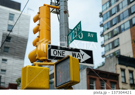 Street signs for West 23 Street and 10 Avenue are displayed above a one way traffic sign in New York City. Surrounding buildings and traffic signals define the urban streetscape. 137304421