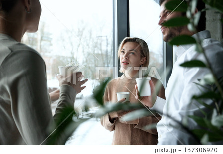 Three people are standing in front of a window, smiling and holding cups 137306920