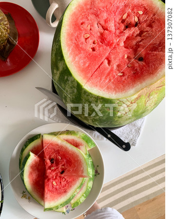 A large watermelon is cut open on a table, revealing its red flesh inside. Slices of the fruit are placed on a plate nearby. This scene shows a casual summer meal setup A large watermelon is cut open on a table, revealing its red flesh inside. Slices of the fruit are placed on a plate nearby. This scene shows a casual summer meal setup 137307082