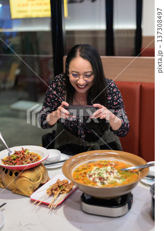 Happy Asian Woman Photographing Spicy Chinese Dish with Smartphone in Restaurant 137308497