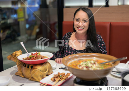 Happy woman sitting at table with hot pot, satay skewers, and stir-fried dish indoors 137308499
