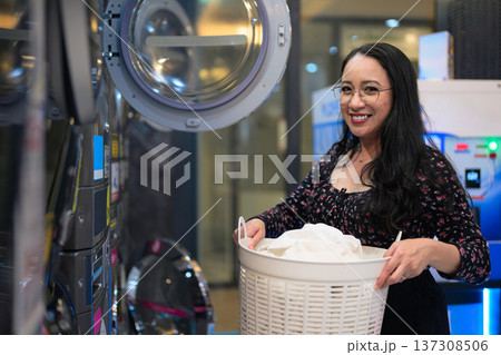 Woman smiling while holding laundry basket near industrial washing machines in public laundromat Woman smiling while holding laundry basket near industrial washing machines in public laundromat 137308506