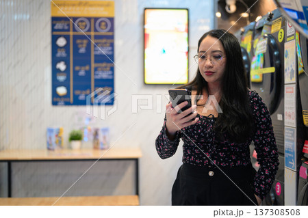 Woman checking mobile phone beside washing machines in modern self-service laundry facility 137308508