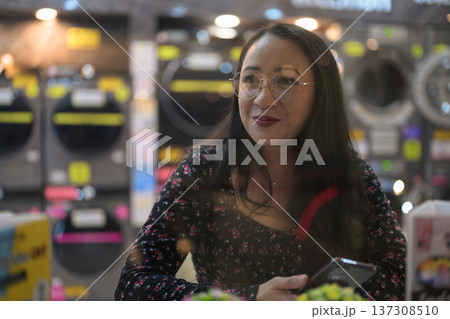 Woman smiling while holding mobile phone inside modern self-service laundromat 137308510