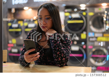 Asian woman looking at mobile phone while sitting inside modern self-service laundromat Asian woman looking at mobile phone while sitting inside modern self-service laundromat 137308512