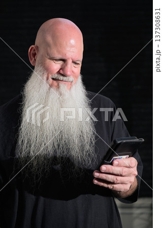 Portrait of a bald happy smiling man with a long white beard using phone against a dark background Portrait of a bald happy smiling man with a long white beard using phone against a dark background 137308631