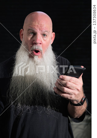 Portrait of a bald man with a long white beard using phone against a dark background 137308634