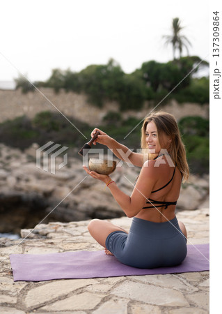 Young woman meditating with a singing bowl on a yoga mat outdoors, practicing sound healing and mindful wellness by the coast Young woman meditating with a singing bowl on a yoga mat outdoors, practicing sound healing and mindful wellness by the coast 137309864