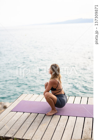 Woman in yoga clothes performing a deep squat pose on a mat. She is meditating by the water next to a wooden pier Woman in yoga clothes performing a deep squat pose on a mat. She is meditating by the water next to a wooden pier 137309871
