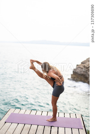 Woman performing a standing balance yoga pose on a mat by the ocean, focusing on flexibility, wellness, and exercise 137309890