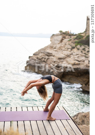 Young woman practicing ustrasana camel pose on a yoga mat overlooking the ocean and rocky cliff, focusing on fitness and wellness 137309891