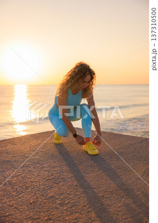 Female runner tying shoelaces on running sneakers before summer workout. Fit young woman at the beach doing sport during vacation Female runner tying shoelaces on running sneakers before summer workout. Fit young woman at the beach doing sport during vacation 137310500