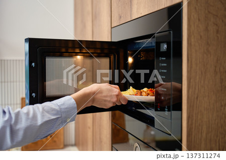 Hand holds plate with cooked food at open built in microwave oven in kitchen cabinet 137311274