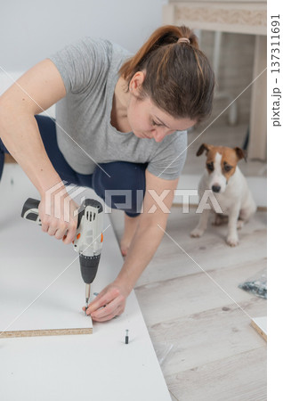A Caucasian woman uses a screwdriver to tighten a screw while assembling a cabinet. A Caucasian woman uses a screwdriver to tighten a screw while assembling a cabinet. 137311691