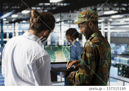 Army officer and IT expert installing server security systems on laptop, control center server room on industrial platform. Military data center engineer and soldier overseeing tactical operations. Army officer and IT expert installing server security systems on laptop, control center server room on industrial platform. Military data center engineer and soldier overseeing tactical operations. 137315190