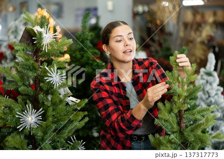 Young female in plaid shirt buying artificial Christmas trees before holidays in a store Young female in plaid shirt buying artificial Christmas trees before holidays in a store 137317773