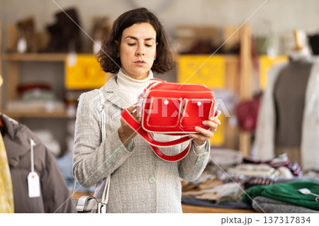 Fashionable female client standing against background of shelves with clothes in modern store and choosing handbag 137317834