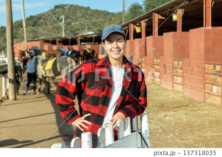 Portrait of young female stable worker smiling at camera during work in horse club on sunny spring day 137318035