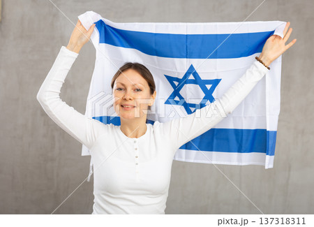 Positive patriotic middle-aged woman holding state flag of Israel against gray wall background indoors Positive patriotic middle-aged woman holding state flag of Israel against gray wall background indoors 137318311