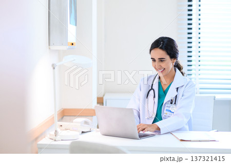 Smiling professional female doctor in white medical coat working with laptop computer, sitting at the table near the window in hospital office, looking at laptop screen. Smiling professional female doctor in white medical coat working with laptop computer, sitting at the table near the window in hospital office, looking at laptop screen. 137321415