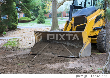 Yellow tractor is digging up dirt in yard surrounded by trees grass on sunny day 137321566