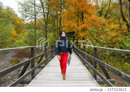Russian woman tourist 50 years old walking on a wooden bridge in autumn forest. Mature female traveler enjoying fall nature and colorful trees. 137322621