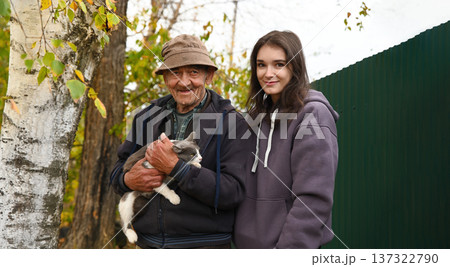 Russian grandfather with his adult granddaughter holding a gray cat outdoors in autumn village garden. Warm authentic rural family moment. 137322790