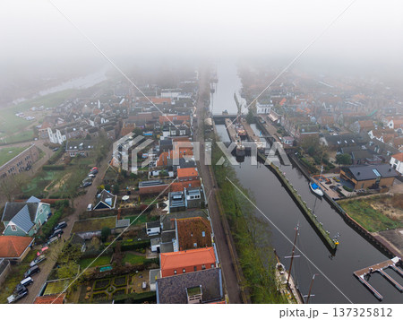 Aerial drone view of a historic Dutch town with a long canal and traditional houses. Foggy morning over the water and rooftops of Muiden. Atmospheric urban landscape in the fog. Aerial drone view of a historic Dutch town with a long canal and traditional houses. Foggy morning over the water and rooftops of Muiden. Atmospheric urban landscape in the fog. 137325812