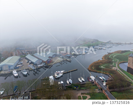 Aerial view of a misty marina in a historic European town. Foggy morning over a harbor with boats, sailboats, and Dutch architecture. Atmospheric drone shot of a quiet winter port. 137325816