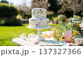 Two tier cake featuring a beautiful hand painted floral design with a bird, set on a decorated table for an outdoor event 137327366