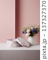 Round floral gift box with lid resting beside a white vase holding a colorful wildflower bouquet on a pink wall backdrop 137327370