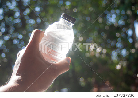 Hand holds a cold plastic water bottle outdoors, glowing in sunlight with soft green bokeh lit sun Hand holds a cold plastic water bottle outdoors, glowing in sunlight with soft green bokeh lit sun 137327830