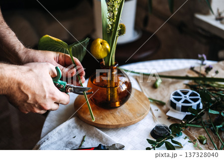 Hands use garden shears to trim green stem near amber vase on table. Floral design course, continuing education for creatives, artisanal training. 137328040