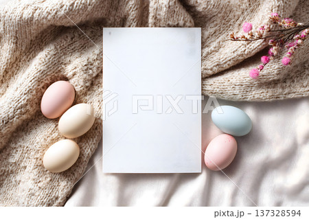 Soft knitted blanket holds white card. Pastel eggs rest beside it gently. Pink blossoms add delicate charm. Natural light highlights textured fabric. Blank card invites your message 137328594