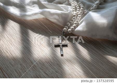 Soft light falls on a silver cross and pearl beads. They rest upon a textured, white fabric. Gentle shadows dance across the wooden surface. The cross hangs low, detailed with engraved lines 137328595