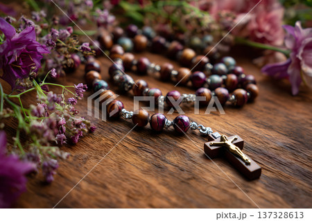 Wooden table holds a beaded rosary. Purple flowers frame the sacred beads. Metal cross gleams beside the rosary. Brown and purple beads display intricate patterns Wooden table holds a beaded rosary. Purple flowers frame the sacred beads. Metal cross gleams beside the rosary. Brown and purple beads display intricate patterns 137328613