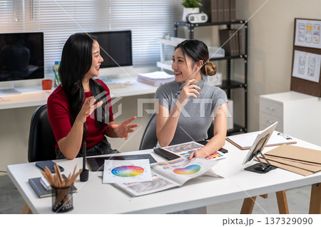 Asian woman holding color chart talking to coworker sitting with working material on table in office 137329090