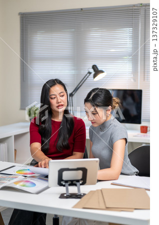 Asian woman explaining color chart to friend or coworker aside tablet at table in home office studio Asian woman explaining color chart to friend or coworker aside tablet at table in home office studio 137329107