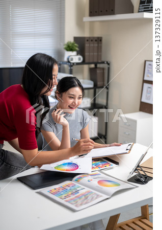 Pretty asian woman and a friend or coworker talking and looking at tablet on table in office studio. 137329115